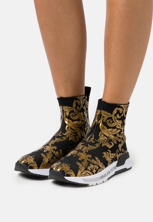 Svarta strumpliknande skor med ett guldornamenterat mönster, texturerad ovandel och vit sula med "Versace Jeans Couture"-branding.