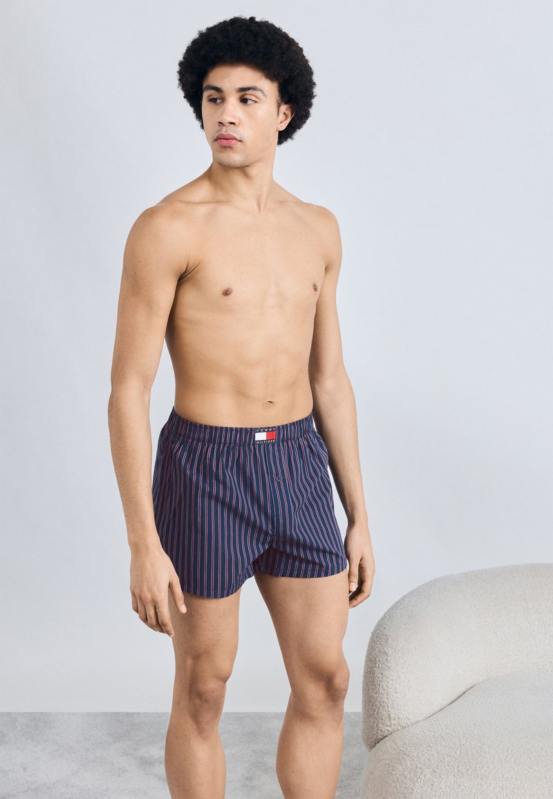 Μακρύβια σορτσάκια σε ναυτικό μπλε με κάθετες κόκκινες και λευκές ρίγες. Ελαστική μέση με μικρή ετικέτα λογότυπου. Ομαλό ύφασμα, άνετη εφαρμογή.