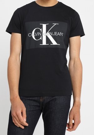 T-shirt noir à manches courtes avec un grand logo "CK" blanc et un logo "Calvin Klein Jeans" gris centré sur la poitrine, porté avec un jean bleu foncé.