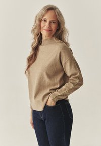 Béžový pletený sveter s vysokým golierom, ozdobený zeleným kontrastným prešívaním na rukávoch, v kombinácii s tmavomodrými džínsami.