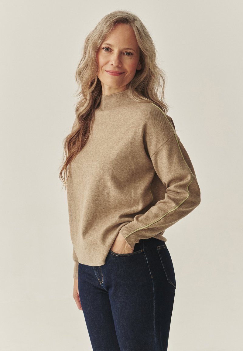 Béžový pletený sveter s vysokým golierom, ozdobený zeleným kontrastným prešívaním na rukávoch, v kombinácii s tmavomodrými džínsami.