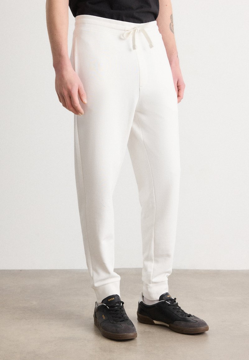Hvide sweatpants med elastisk talje og snøre, tapered ben og sidelommer, parret med sorte sportssko med gummisål.