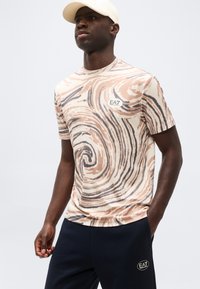 Άνδρας που φοράει μπεζ και μαύρο μπλουζάκι με σχέδιο στροβιλισμού και λογότυπο EA7, μαύρο παντελόνι με το ίδιο λογότυπο και μπεζ καπέλο, στέκεται μπροστά σε απλό φόντο.