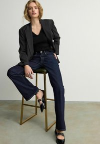Blazer noir à rayures, top noir avec décolleté en V, jeans en denim droits bleu foncé et chaussures noires avec détails en boucle, assis sur un tabouret en métal.