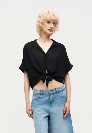 Ξανθό άτομο με μια χαλαρή μαύρη πουκάμισο με κουμπιά, δεμένη στη μέση, και ανοιχτό μπλε τζιν με ψηλή μέση, σε απλό φόντο.