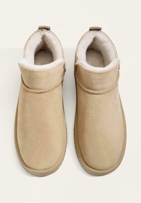 Para beżowych zamszowych kapci wsuwanych z puchowym białym wnętrzem i okrągłymi noskami, widocznych z góry na jasnym tle.