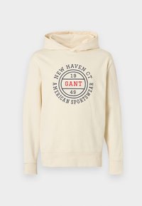 Φούτερ με κουκούλα σε εκρού χρώμα από μαλακό υλικό, με κυκλικό λογότυπο που αναγράφει "GANT" σε κόκκινο και "NEW HAVEN CT 1949 AMERICAN SPORTSWEAR" σε μπλε σκούρο.