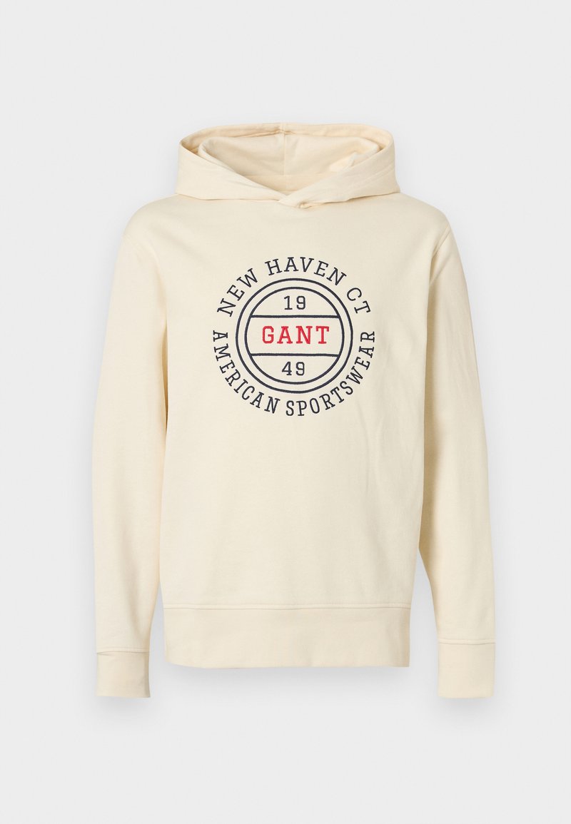 Sweat à capuche crème en matière douce, avec un logo circulaire affichant "GANT" en rouge et "NEW HAVEN CT 1949 AMERICAN SPORTSWEAR" en bleu marine.