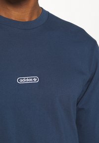 Námořnická modrá tričko s dlouhým rukávem vyrobené z bavlny, s bílým logem Adidas ve kulaté badge designu na hrudi.