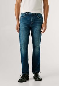 Vaqueros de mezclilla azul oscuro con corte recto, que cuentan con bolsillos delanteros, detalles de costura y una textura ligeramente descolorida. Combinados con zapatos negros.