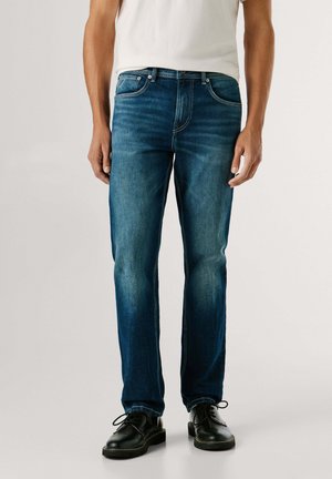 Donkerblauwe spijkerbroek met een rechte pasvorm, voorzien van voorzakken, stikseldetails en een licht vervaagde textuur. Draagt zwarte schoenen.