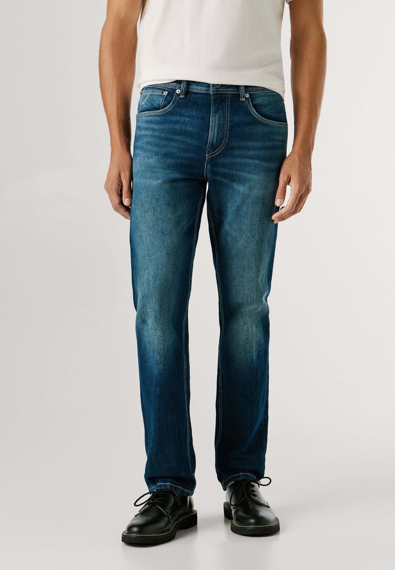 Vaqueros de mezclilla azul oscuro con corte recto, que cuentan con bolsillos delanteros, detalles de costura y una textura ligeramente descolorida. Combinados con zapatos negros.