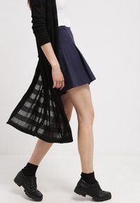 Cardigan aperto nero in maglia, top fitted bianco, gonna plissettata blu navy, calzini neri alla caviglia e scarpe lacciate nere chunky. Sfondo con terreno pianeggiante.