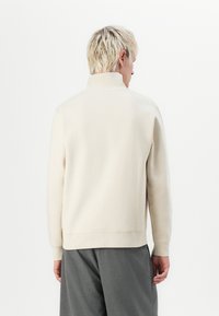 Beige yliopistopaita korkealla kauluksella, pitkät hihat ja ribatut hihansuut. Pehmeä rakenne, minimalistinen muotoilu ja rento istuvuus.