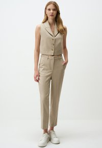 Beige ärmellose cropped Blazer mit vier Knöpfen, kombiniert mit passenden maßgeschneiderten Hosen. Die Schuhe sind weiße Sneakers. Glatte Stofftextur.