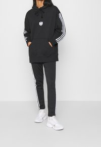 Black hoodie with a front pocket and embroidered logo, paired with black pants featuring white side stripes, finished with white sneakers.
