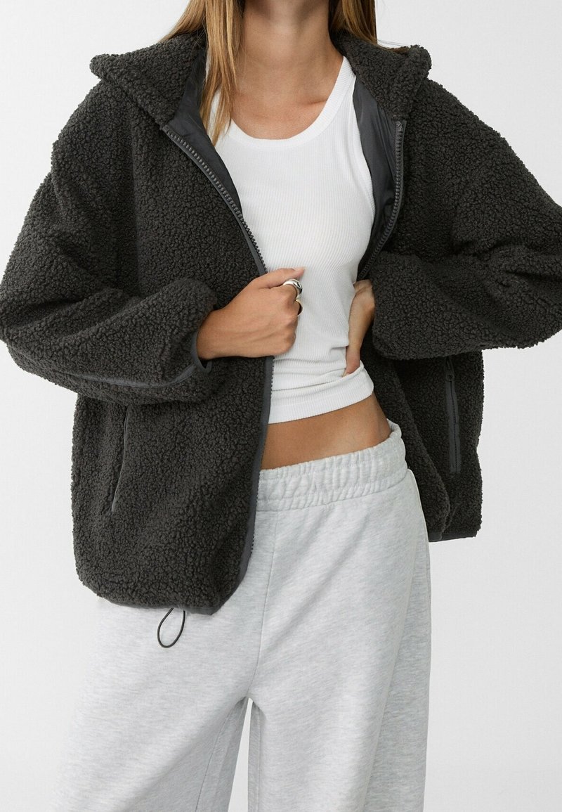 Grijze fleece jas met een zachte, gestructureerde afwerking, met ritssluiting aan de voorkant en zijzakken. Gecombineerd met een witte, aansluitende shirt en grijze joggingbroek.