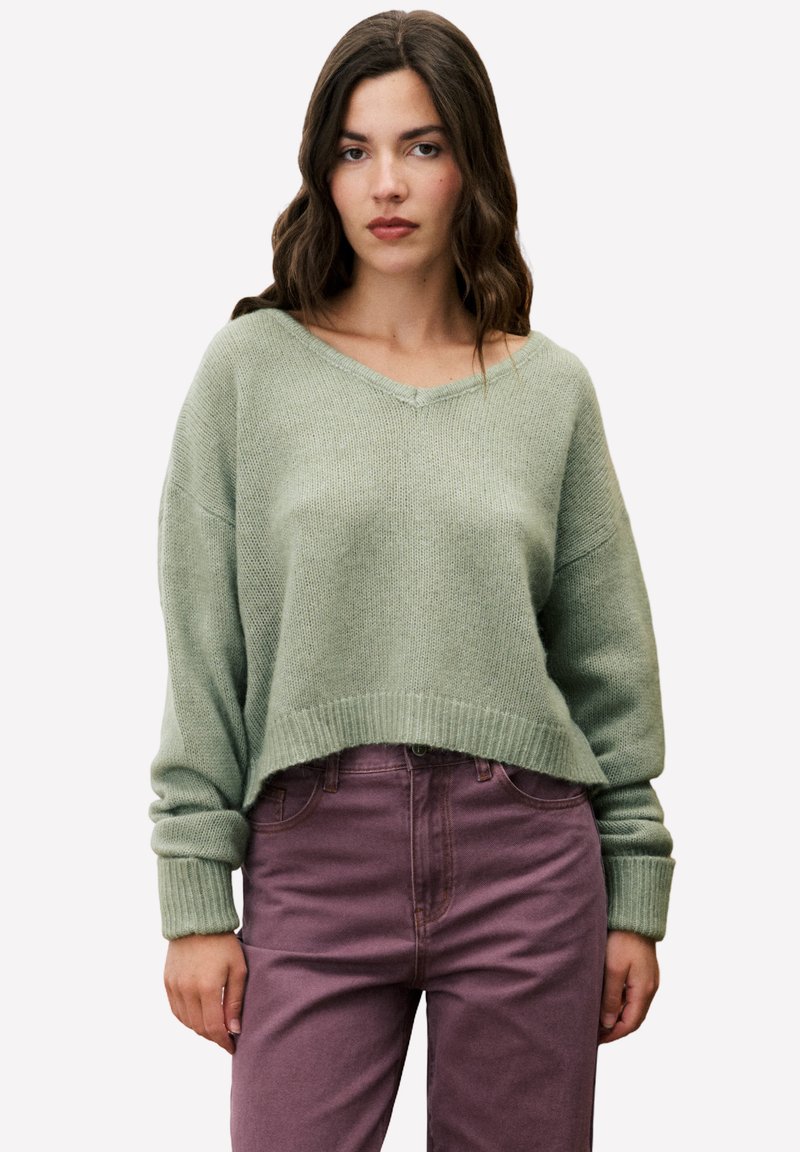 Grüner Strickpullover mit V-Ausschnitt, weiten Ärmeln und geripptem Saum. Kombiniert mit lila Jeans f�ür einen lässigen Look. Weiche Textur.