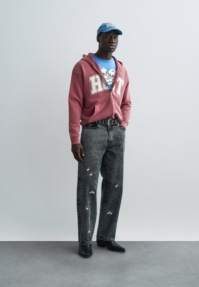 Sarkans вverters džemperis ar tekstu “HOT”, zils grafiskais t-krekls, nodilušas pelēkas džinsa bikses ar baltiem zaķu rakstiem un melnas smailas kurpes.