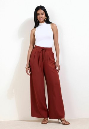 Femme aux longs cheveux noirs portant un haut blanc sans manches et un pantalon large taille haute couleur rouille avec ceinture à nouer, debout contre un mur blanc.