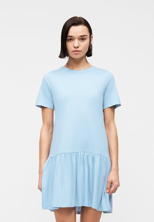 Młoda kobieta o krótkich ciemnych włosach w jasnoniebieskiej sukience z krótkim rękawem i marszczoną falbaną u dołu, stojąca na jednolitym tle.