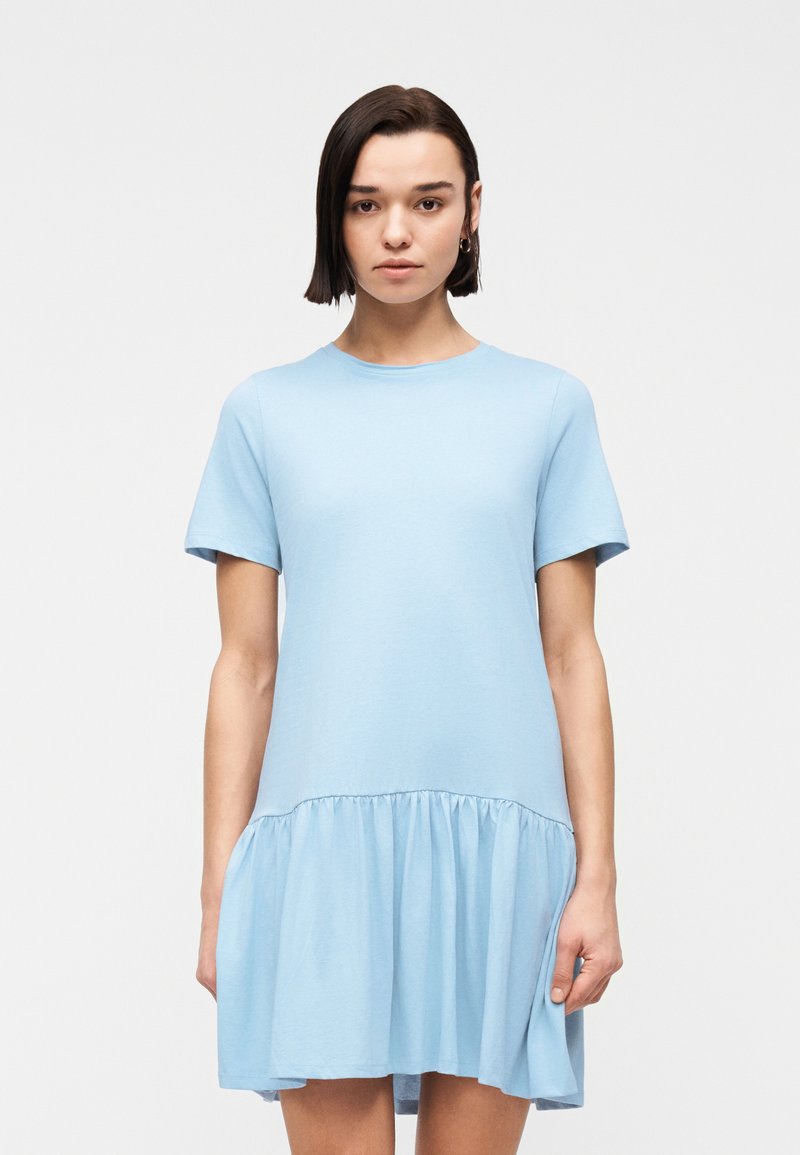 Νεαρή γυναίκα με κοντά σκουρόχρωμα μαλλιά, που φοράει ανοιχτό μπλε φόρεμα με κοντά μανίκια και βολάν στο τελείωμα της φούστας, στέκεται μπροστά σε απλό φόντο.