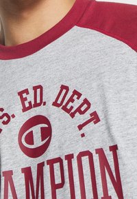 Camiseta de algodón gris con mangas raglán rojas, que presenta un atrevido texto en blanco y rojo que dice "CHAMPION" y "U.S. ED. DEPT." en la parte delantera.