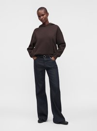Personne portant un sweat à capuche marron foncé et un jean bleu foncé à jambes larges avec des chaussures noires, debout les mains dans les poches sur un fond uni.