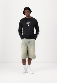 Svart langermet skjorte med grafisk trykk, lyse denimshorts, hvite sneakers og en svart bøttehatt, som står mot en enkel bakgrunn.