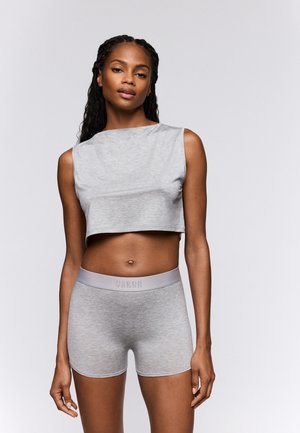 Vrouw die een lichtgrijze, mouwloze cropped top en bijpassende hoge taille shorts draagt, staand tegen een effen witte achtergrond.