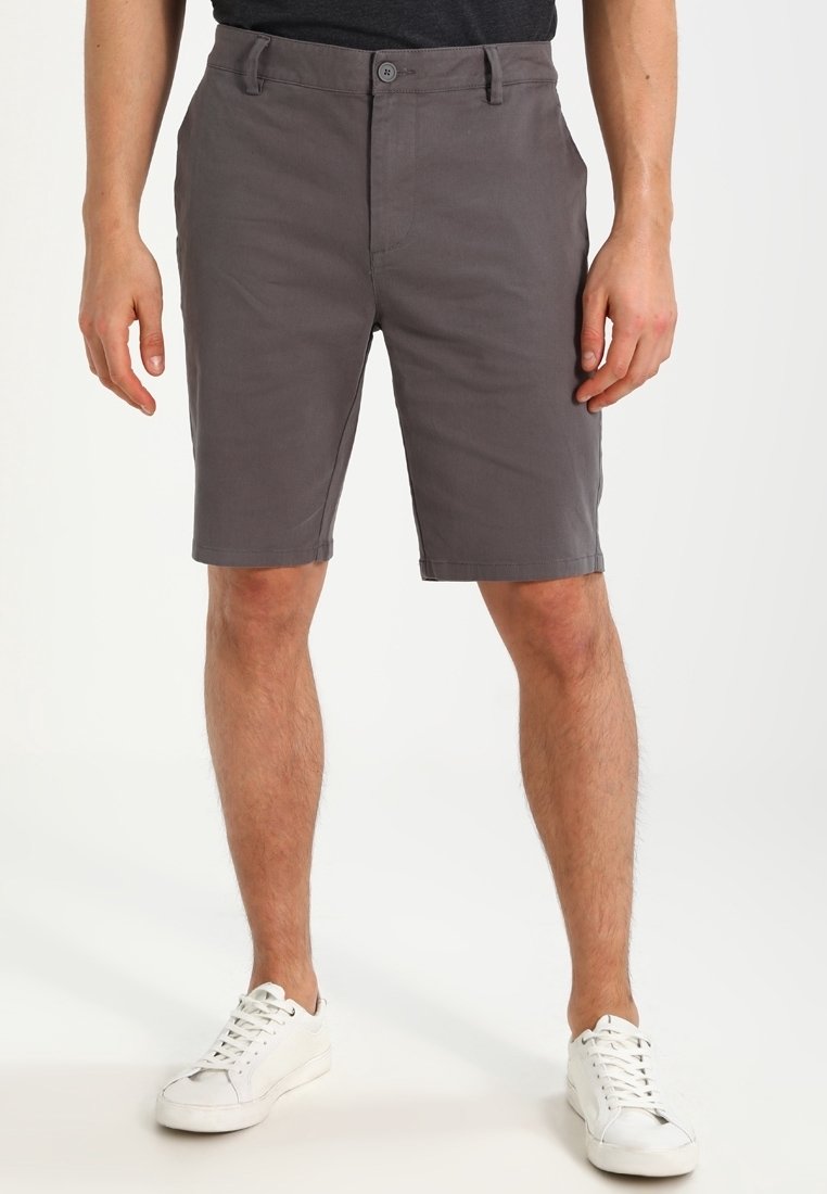 Homme portant un short gris foncé jusqu'aux genoux et des baskets blanches, debout avec les mains détendues le long du corps sur un fond blanc.