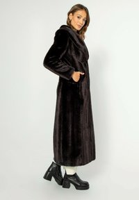 Černý kabát z umělé kožešiny dlouhého střihu s klopovým límcem a bočními kapsami. Model má na sobě robustní černé boty na podpatku.