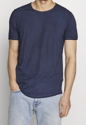 Homme portant un t-shirt uni bleu marine à manches courtes et un jean bleu clair, une main dans la poche, sur fond blanc.