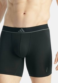 Torse masculin portant un boxer noir Adidas avec une ceinture grise et le logo Aeroready sur la cuisse gauche, sur un fond uni.