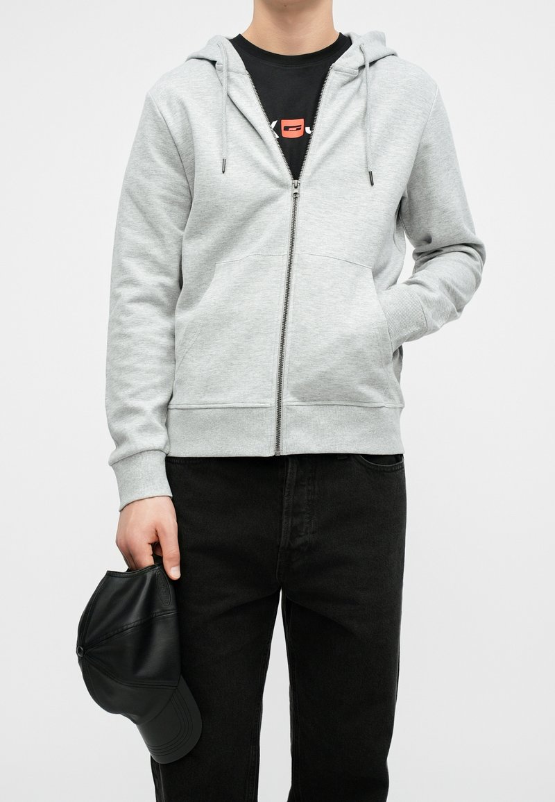 Personne portant un sweat à capuche gris clair zippé par-dessus un t-shirt noir, pantalon noir, tenant une casquette de baseball en cuir noir à côté d’elle.