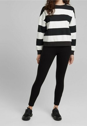 Kvinde iført en sort og hvid langærmet top med brede striber, sorte leggings og sorte sneakers, stående foran en neutral baggrund.