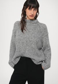 Suéter gris de punto con cuello alto, corte holgado y puños acanalados. Tela texturizada con una apariencia acogedora, combinado con pantalones negros.