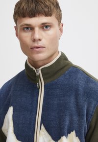 Giovane uomo con capelli corti che indossa una giacca in pile con zip, caratterizzata da un motivo a montagna blu e bianco e un colletto verde oliva.