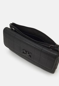Svart läderclutch med quiltat mönster, upphöjd CK-logotyp, dragkedjestängning upptill och slät textur. Kompakt rektangulär form.