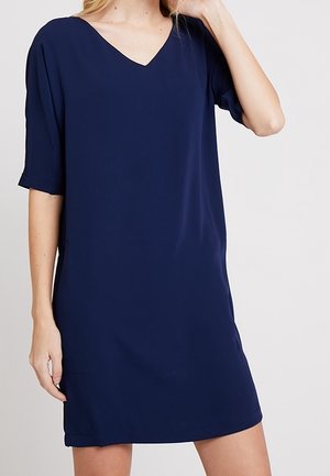 Femme portant une robe simple bleu marine, manches courtes, col en V, longueur genou, debout devant un fond uni.