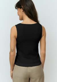 Svart ärmlös topp i en texturerad tyg, med rundad halsringning och en kroppsnära design, ihop med ljusbeige byxor.