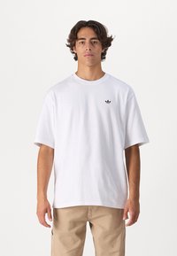 T-shirt bianco in cotone con maniche corte e scollatura rotonda, caratterizzato da un piccolo logo nero sul lato sinistro del petto. Texture liscia, vestibilità rilassata.