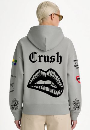 Personne aux cheveux blonds courts portant des lunettes de soleil et un sweat à capuche gris avec des lèvres noires et le texte "Crush" dans le dos ainsi que divers graphiques sur les manches.