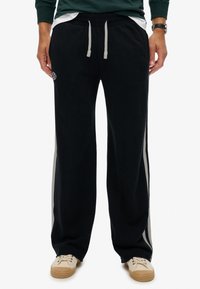 Man met zwarte wijde sweatpants met witte zijstrepen, grijze trekkoorden, een groene sweatshirt en beige sneakers.