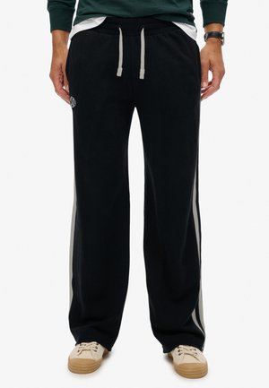 Mand iført sorte wide-leg sweatpants med hvide sidelister, grå snoretræk, en grøn sweatshirt og beige sneakers.
