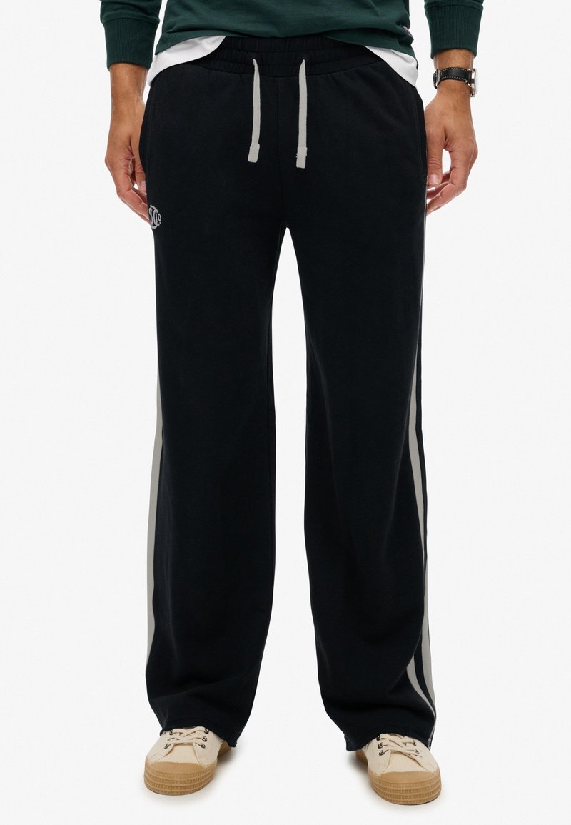Man met zwarte wijde sweatpants met witte zijstrepen, grijze trekkoorden, een groene sweatshirt en beige sneakers.