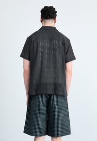 Svart kortermet skjorte i åpen strikk med et teksturert mønster, kombinert med knelengde rutete shorts i mørkegrønn og svart.