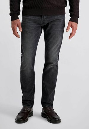 Mann mit abgetragenen schwarzen Jeans, einem dunklen Strickpullover und dunkelbraunen Lederloafers, der vor einem schlichten Hintergrund steht.