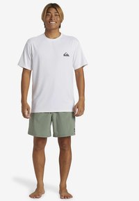 Valge lühikeste varrukatega T-särk, millel on rinnal väike must logo, sobitatud heleroheliste lühikeste pükstega. Puuvillane tekstuur, vabaaja lõige.