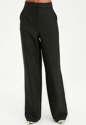 Zwarte, hoog uitgesneden broek met wijde pijpen gedragen door een persoon, gecombineerd met witte schoenen met een zwart bloemmotief.